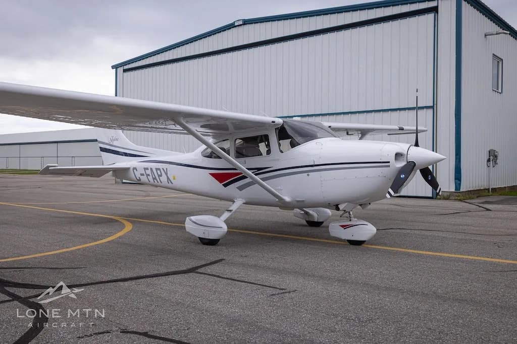 A white Cessna 182 Skylane with grey and blue stripes, registration C-FAPX, parked on an asphalt ramp with a hangar in the background.