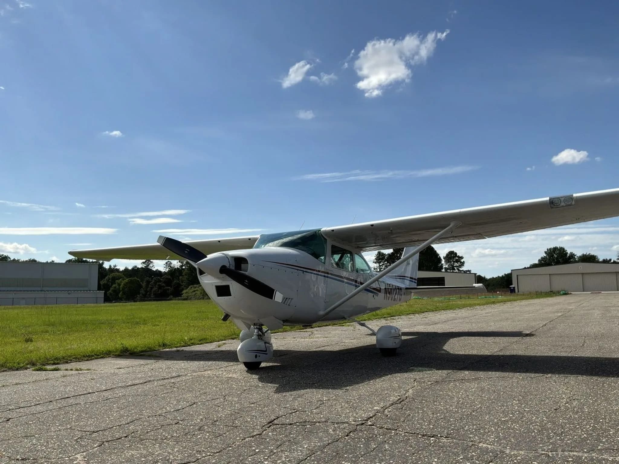 A white Cessna 182N Skylane with red and blue stripes, registration N9027G, parked on an asphalt ramp under a blue sky.