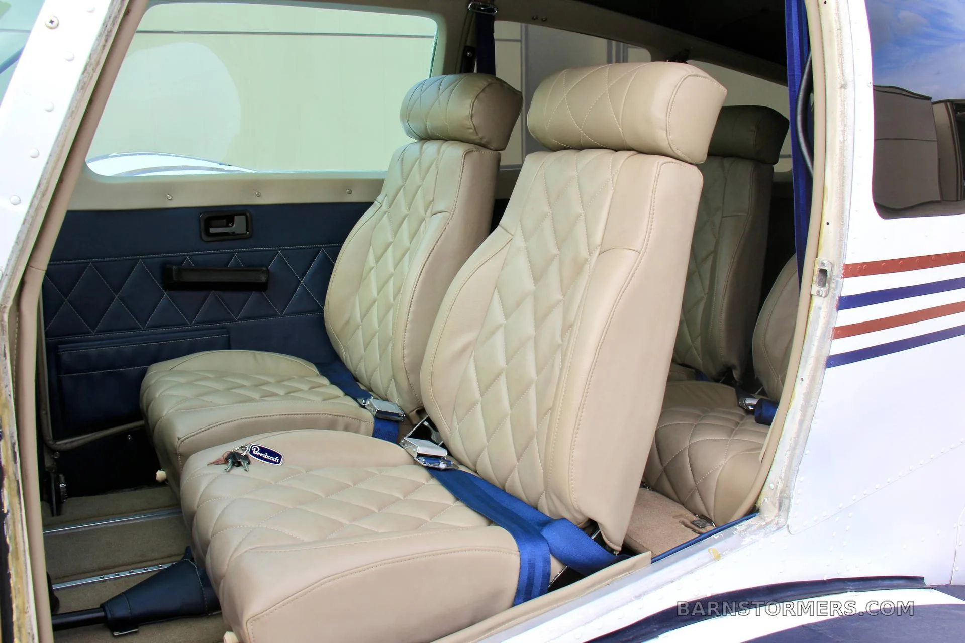 The interior cabin of a small aircraft, showing light tan quilted leather seats with blue seatbelts.