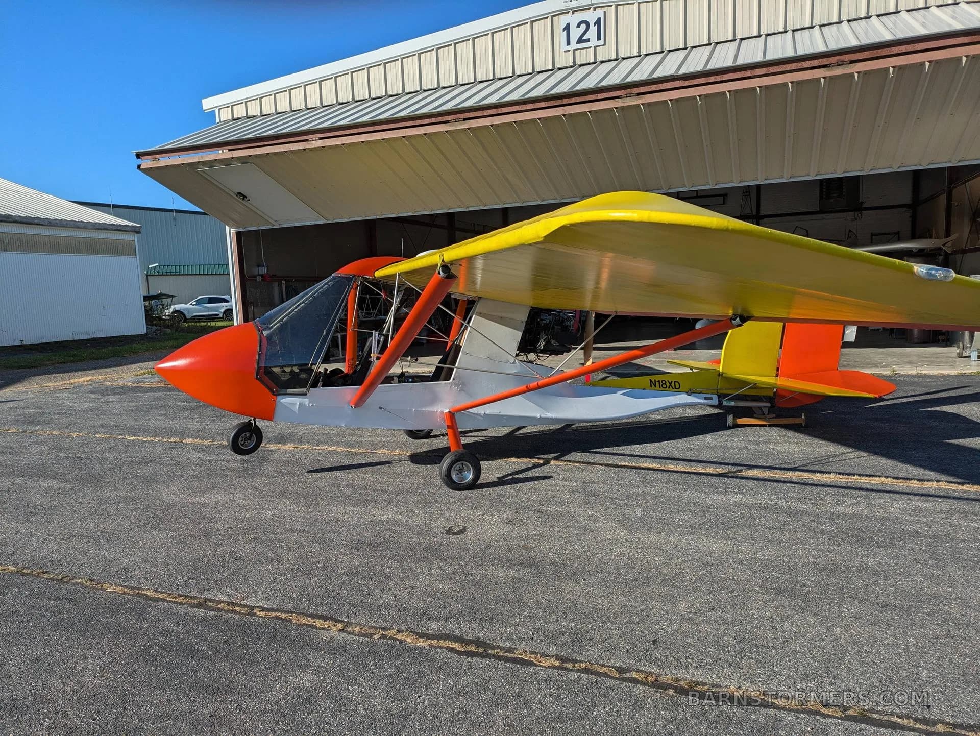 A side view of a red and yellow ultralight aircraft with registration N18XD parked outside a hangar.
