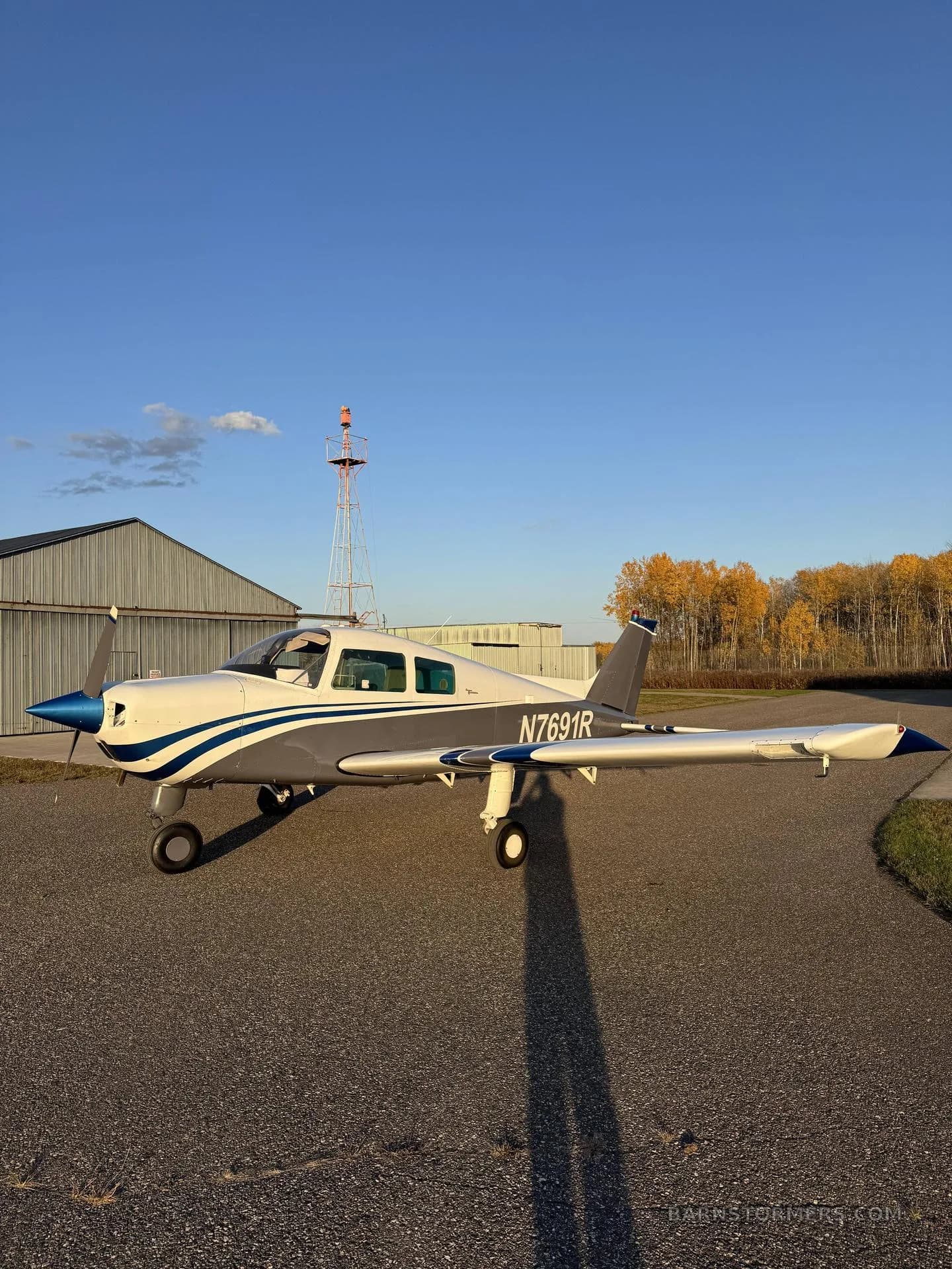 A white and blue single-engine aircraft (N7691R) parked on an asphalt ramp with an airport beacon and autumn trees in the background.