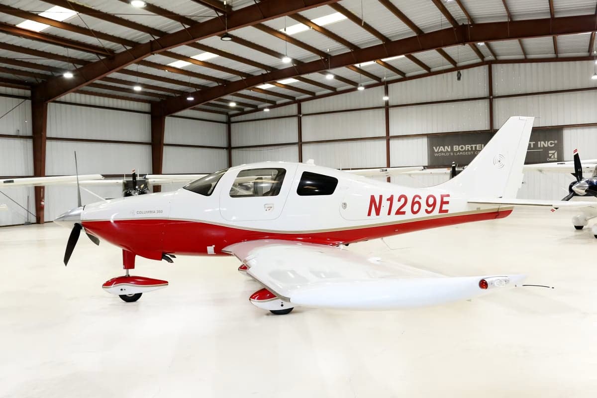 A Columbia 350 aircraft with red and white paint scheme is parked in a hangar.