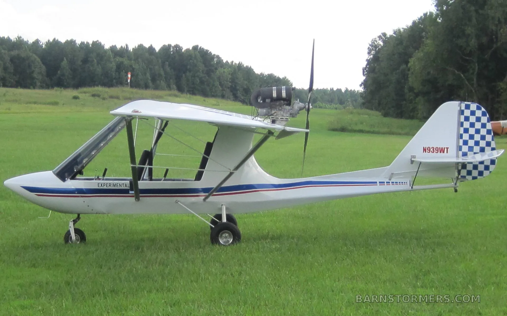 A white experimental aircraft with blue and red stripes and a checkered tail, parked on a grassy airfield.