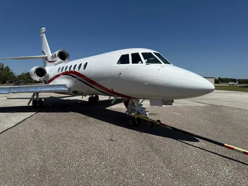 A white Dassault Falcon 900EX business jet with red and black stripes on the tarmac, connected to a tow bar, under a clear blue sky.