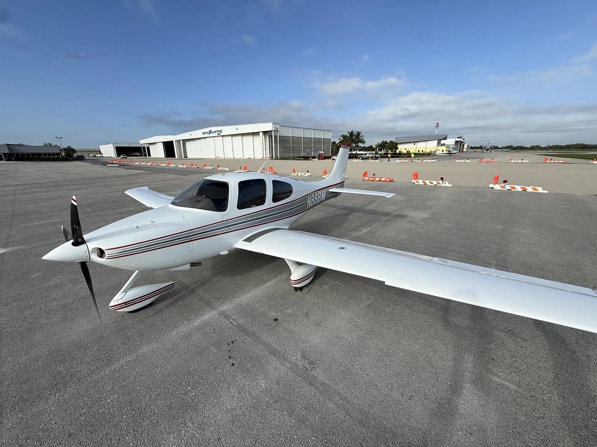 A white Cirrus SR22 aircraft with red and gray stripes is parked on an airport tarmac under a blue sky.