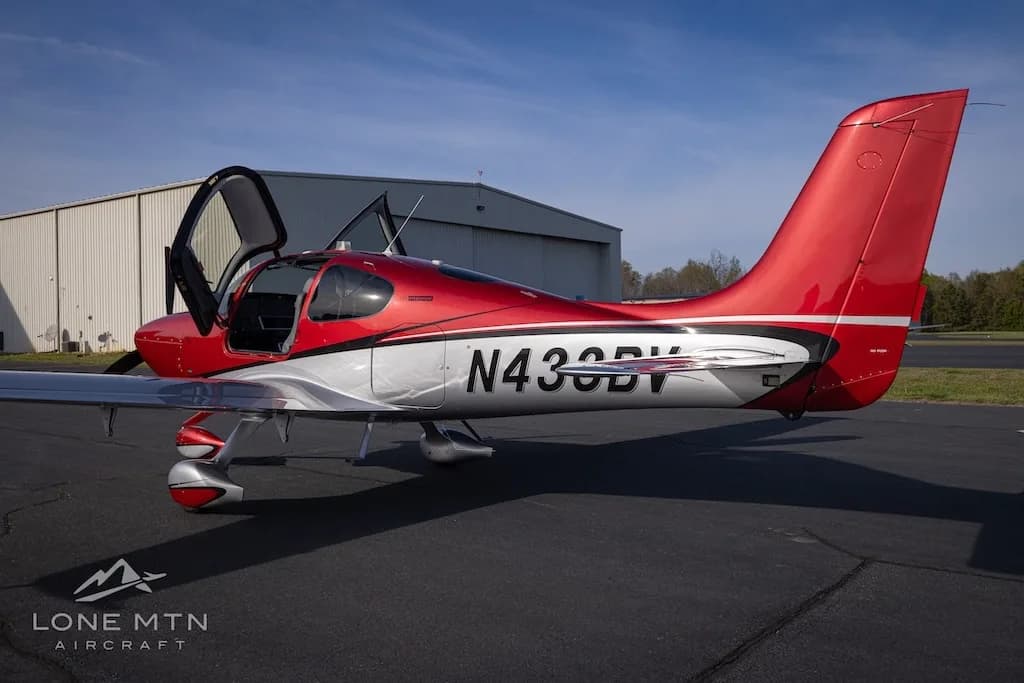 A three-quarter rear view of the red and silver Cirrus SR22 aircraft N433BV with its doors open, parked in front of a hangar.