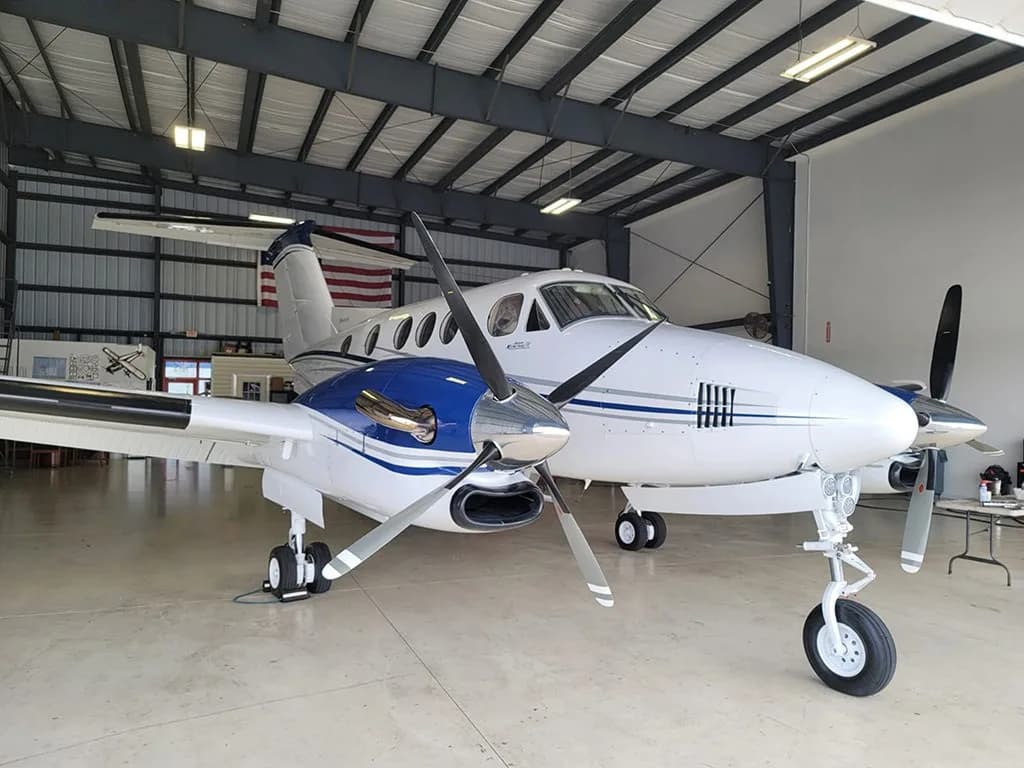 A white and blue twin-engine turboprop aircraft with its nose facing right, parked inside a hangar with an American flag in the background.
