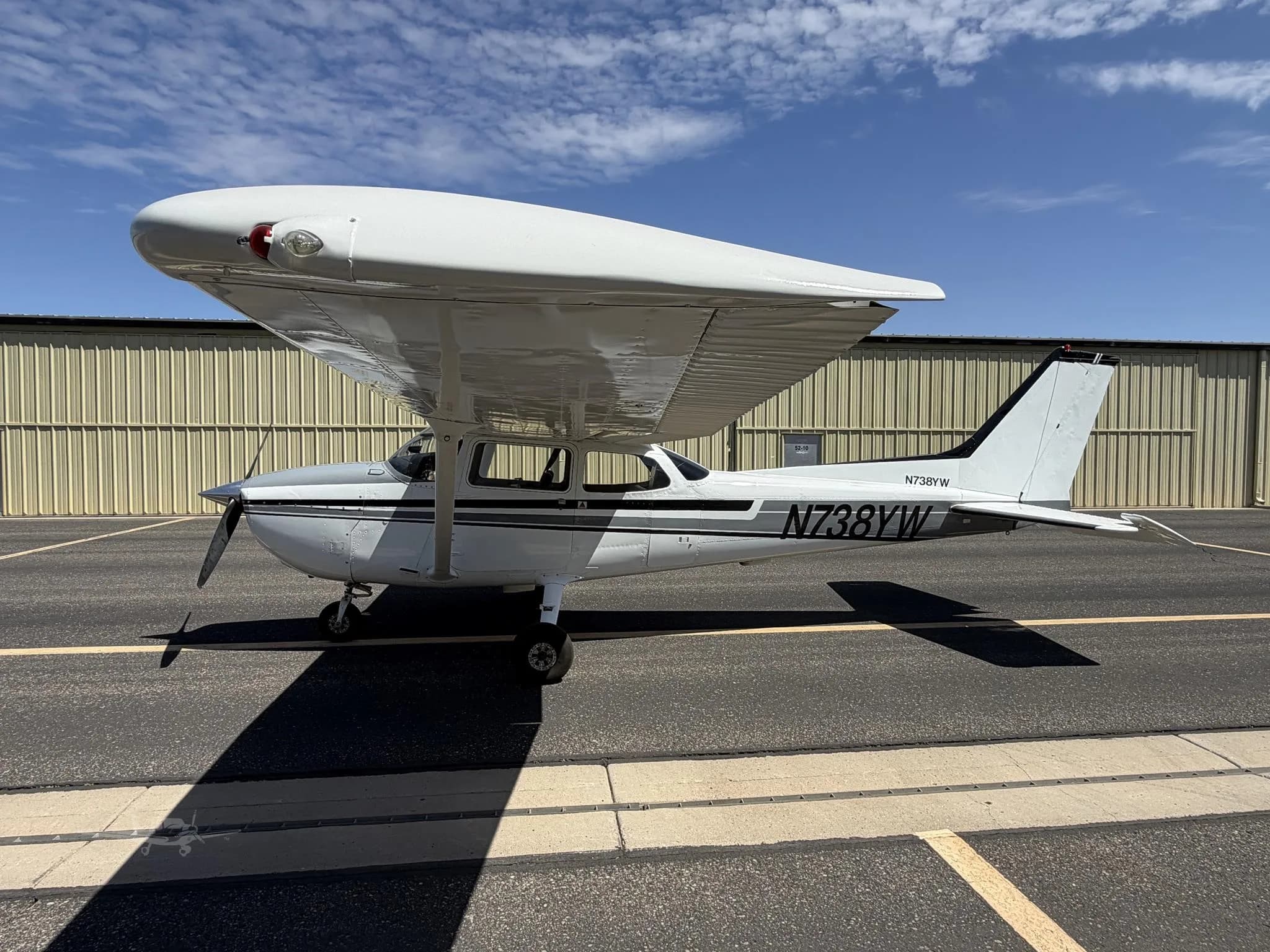 A white high-wing single-engine propeller aircraft with black and grey stripes is parked on an asphalt tarmac next to a hangar under a blue sky.