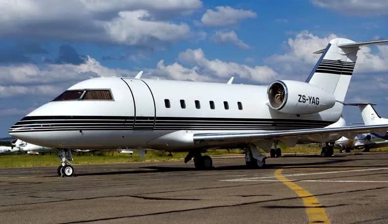 A white Bombardier Challenger business jet with black stripes and registration ZS-YAG on a tarmac under a cloudy sky.
