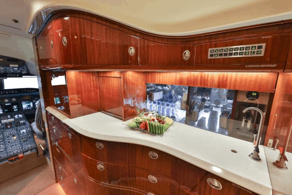 A luxurious aircraft galley with polished wood cabinetry, a white countertop, and a fruit platter, with the cockpit visible in the background.