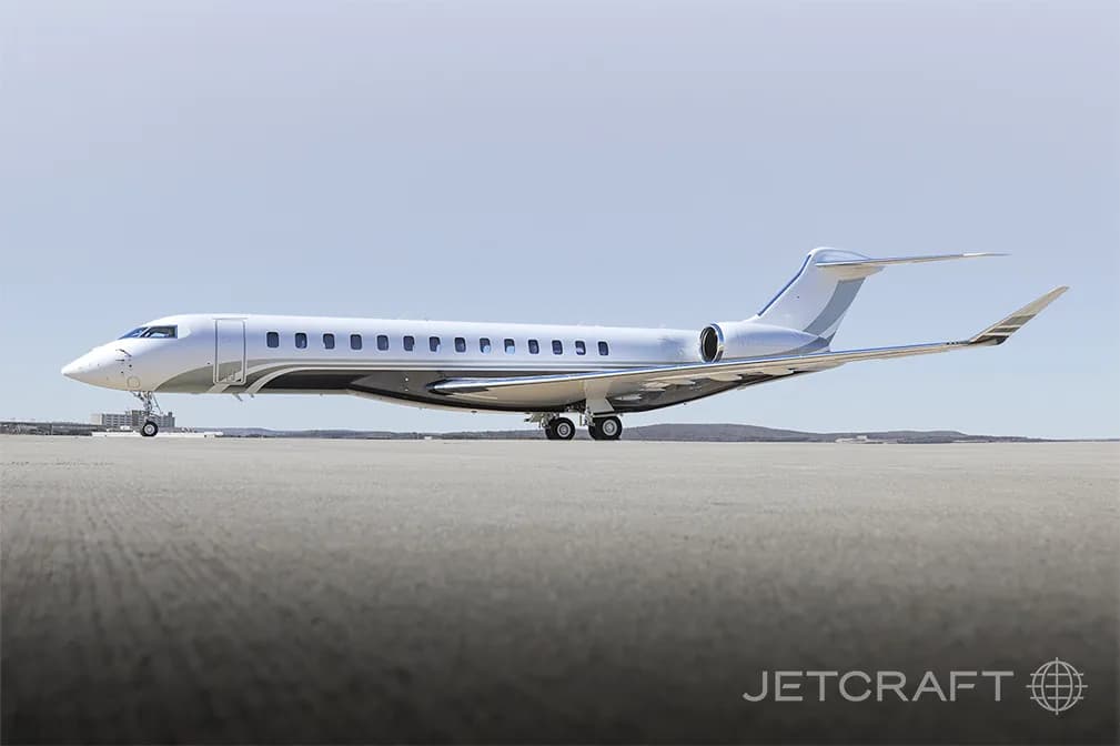 A white and grey Bombardier Global 7500 business jet parked on an airfield under a clear sky.