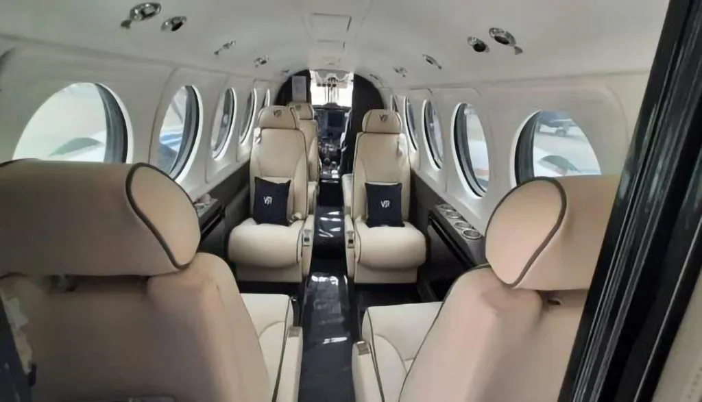A view down the aisle of an aircraft cabin shows light beige leather seats with black pillows and small round windows.