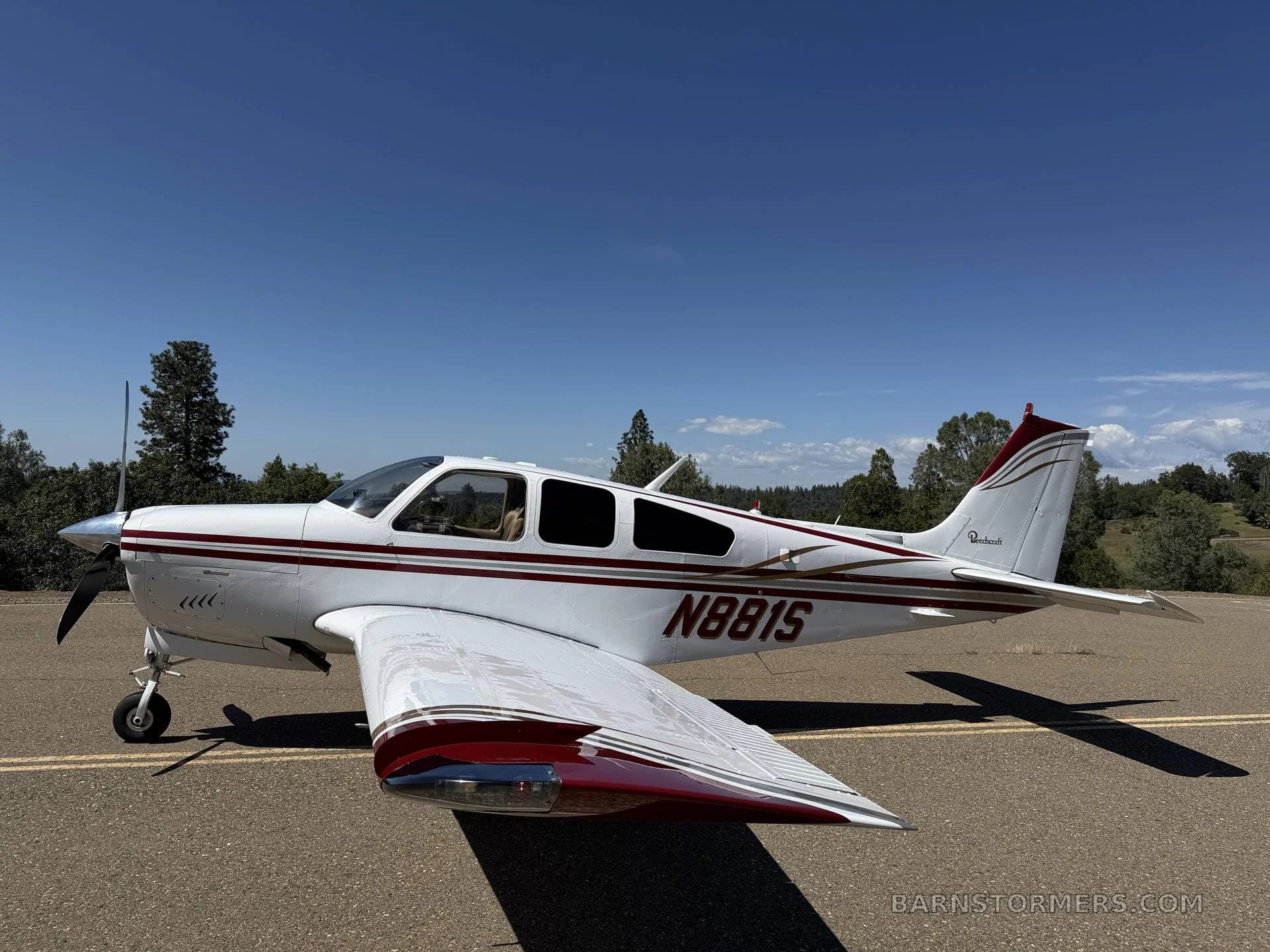 A white Beechcraft Bonanza N881S with red and gold stripes parked on an asphalt surface, viewed from the rear-left.