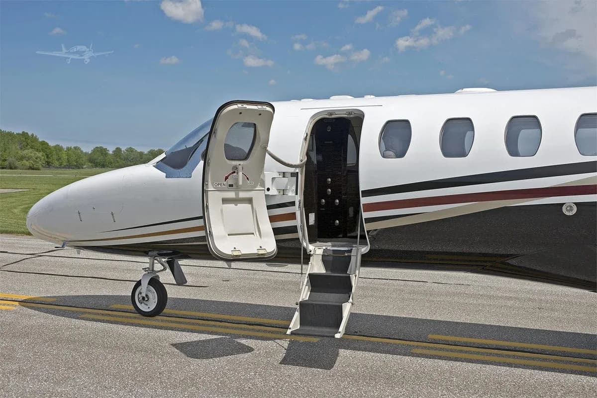 The front section of a white and black business jet with its main entry door open and stairs extended on a sunny tarmac.