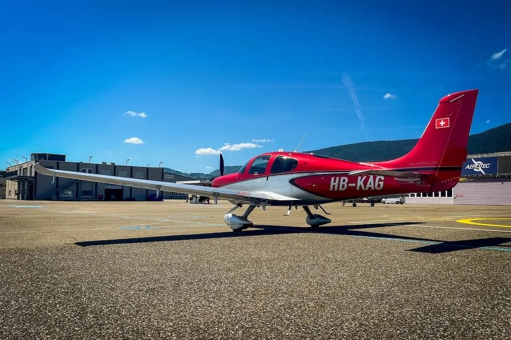 Side view of a red and silver Cirrus SR22 aircraft parked on an airfield with hangars and mountains in the distance.
