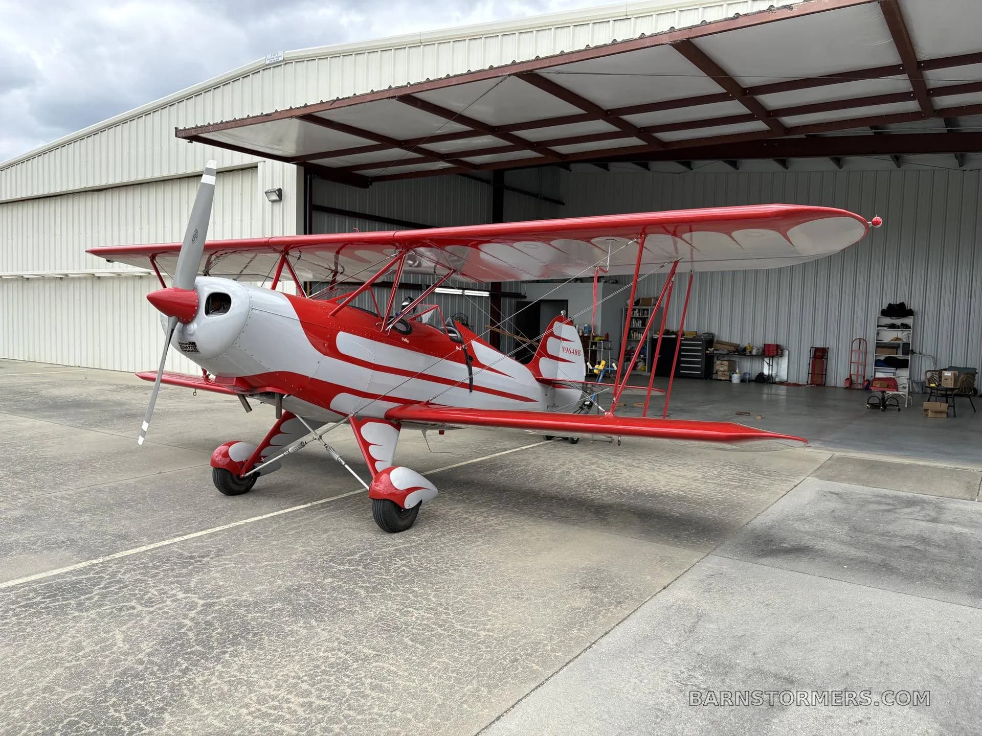 A red and white biplane with a radial engine is parked on concrete in front of an open hangar.