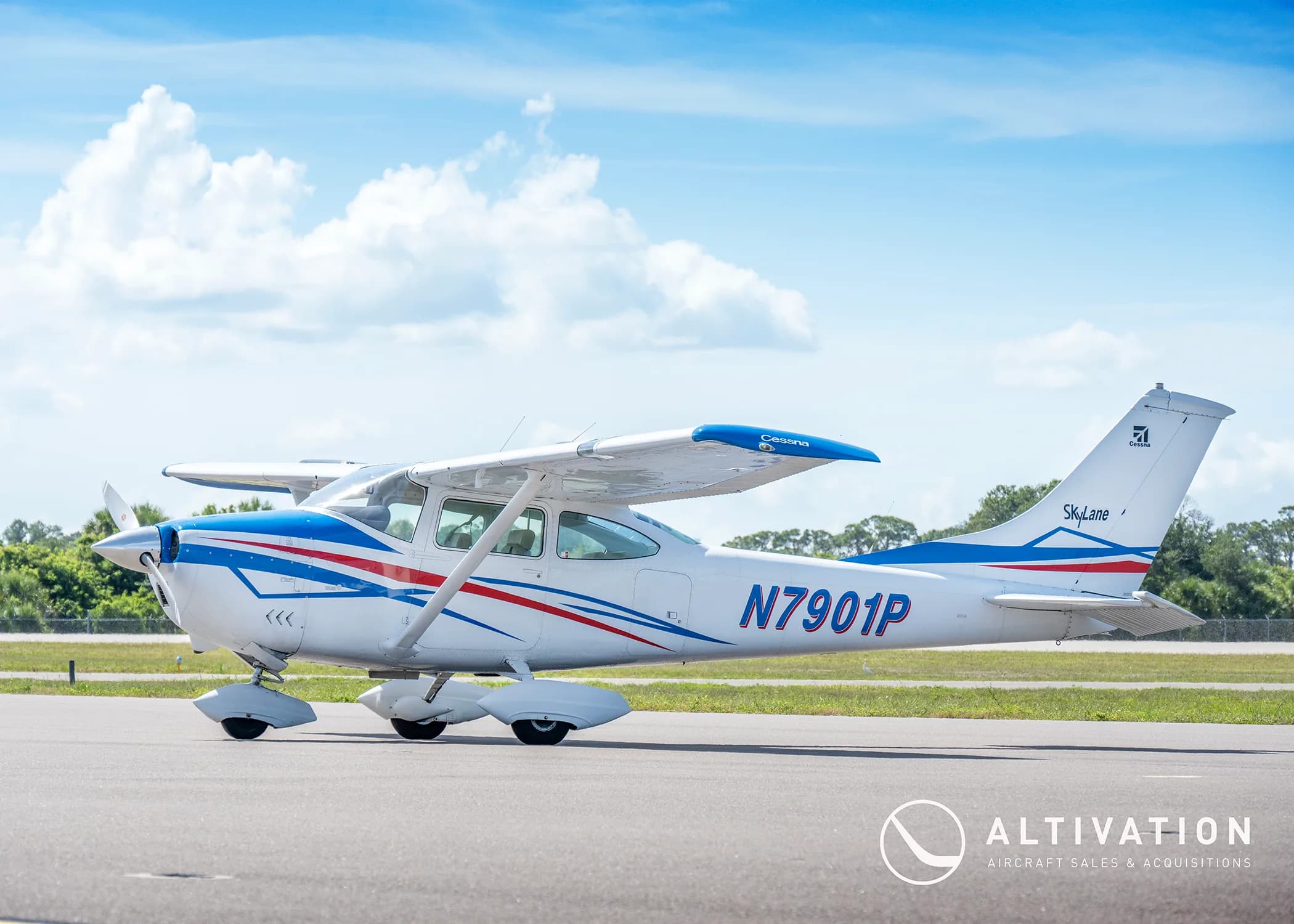 A white Cessna 182 Skylane with blue and red stripes parked on a tarmac with trees in the background.