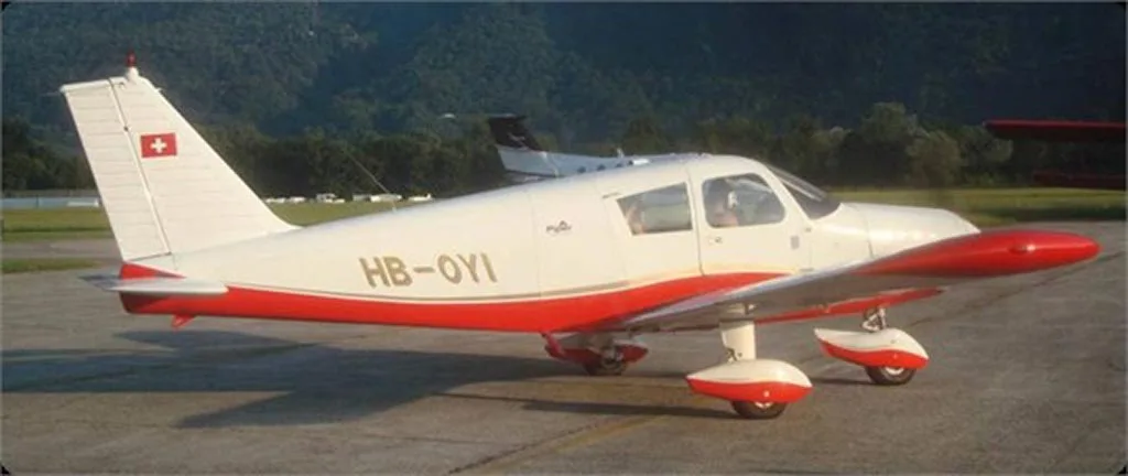 A white and red Piper Cherokee aircraft (HB-OYI) parked on an airfield with a forested mountain and another aircraft in the background.