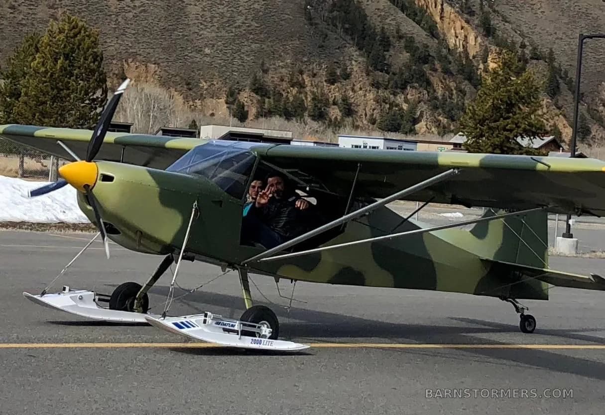 A camouflage-painted light aircraft with skis attached to its wheels has two people visible in the cockpit, parked on an asphalt surface near a mountain.