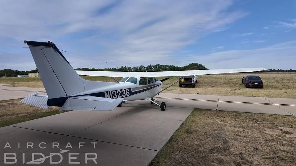 A rear-side view of a white and blue Cessna 172 Skyhawk (N13236) on a taxiway with cars in the distance.