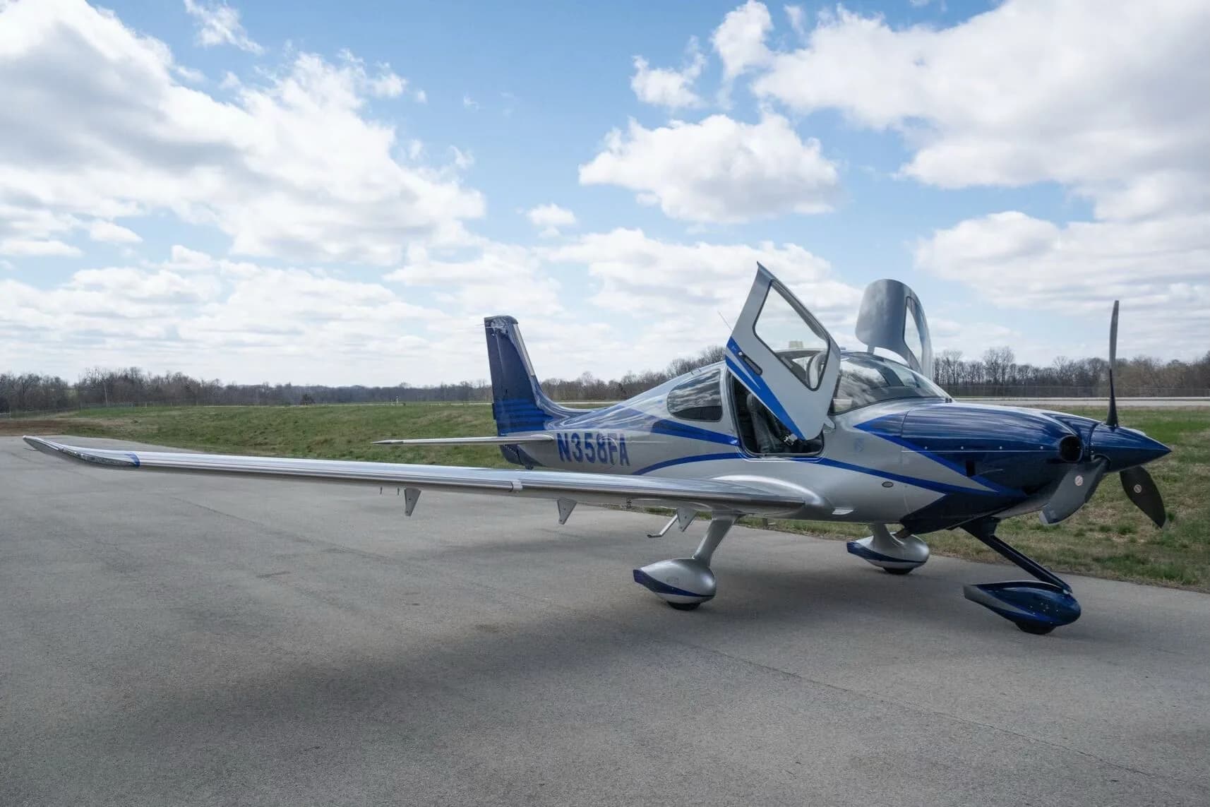 A silver and blue Cirrus SR22T aircraft, registration N358FA, with its gull-wing doors open, parked on an airfield.