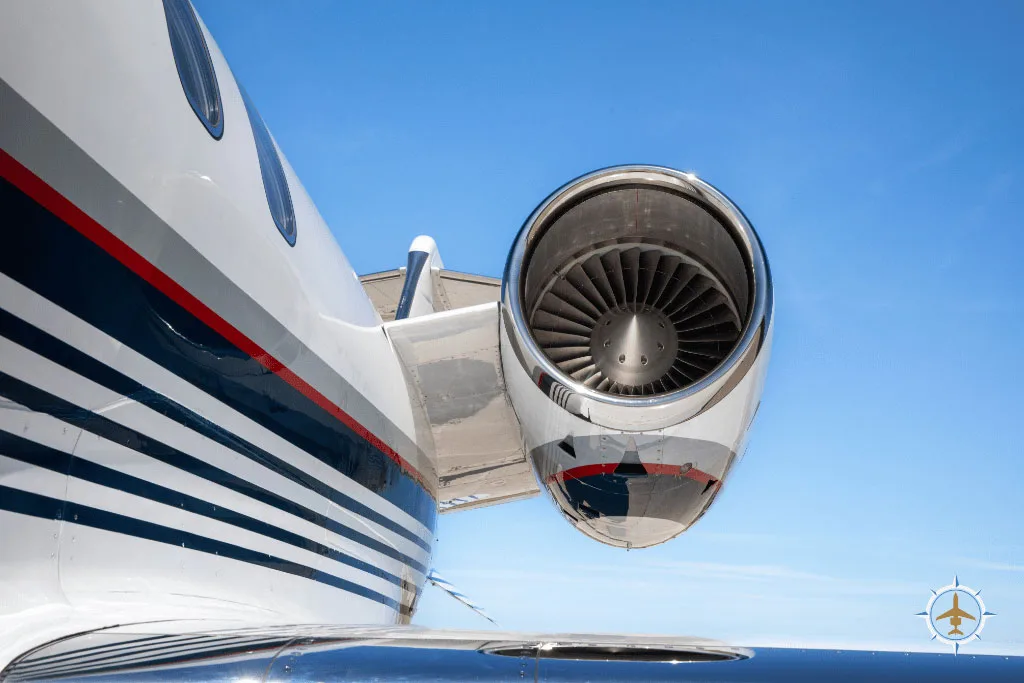 A close-up view of the jet engine and rear fuselage of a white business jet with blue and red stripes against a clear blue sky.