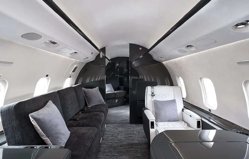 The spacious interior cabin of a private jet, showing a dark grey sofa on the left and a white quilted seat on the right.