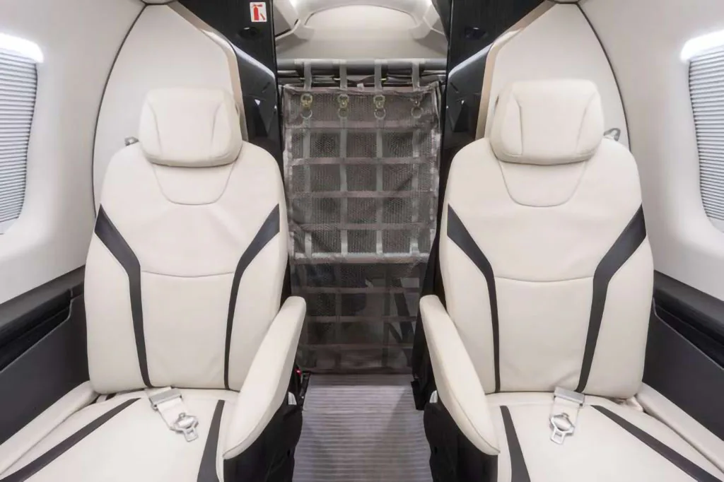 Two white leather seats with black accents are visible in the aircraft cabin, with a cargo net partition behind them.
