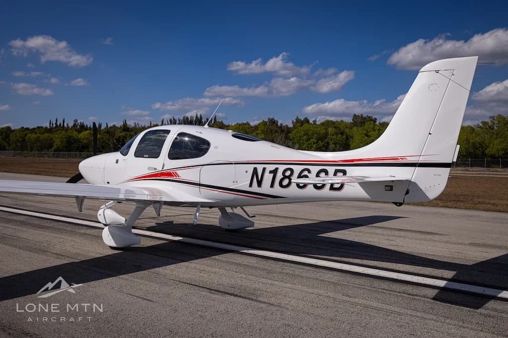 A rear-side view of the white N186SB single-engine aircraft with red and black accent stripes, parked on an airfield.