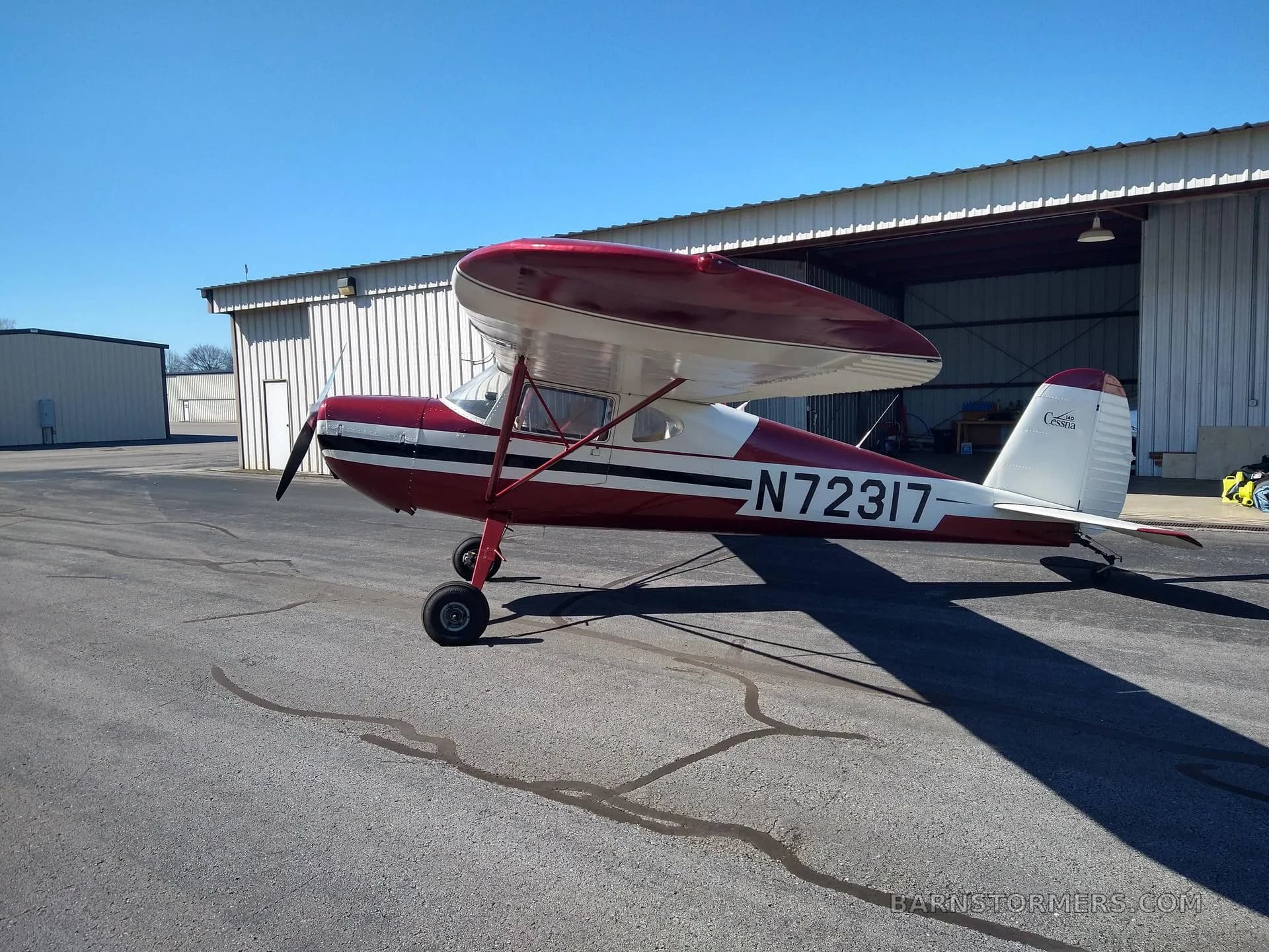 A side view of a red and white Cessna 140 aircraft (N72317) with a high wing, parked on an asphalt tarmac.
