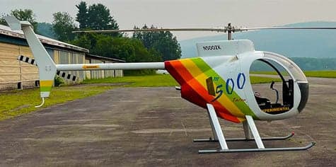 A side view of the N500ZK helicopter with a rainbow paint scheme on the tail, parked on pavement.
