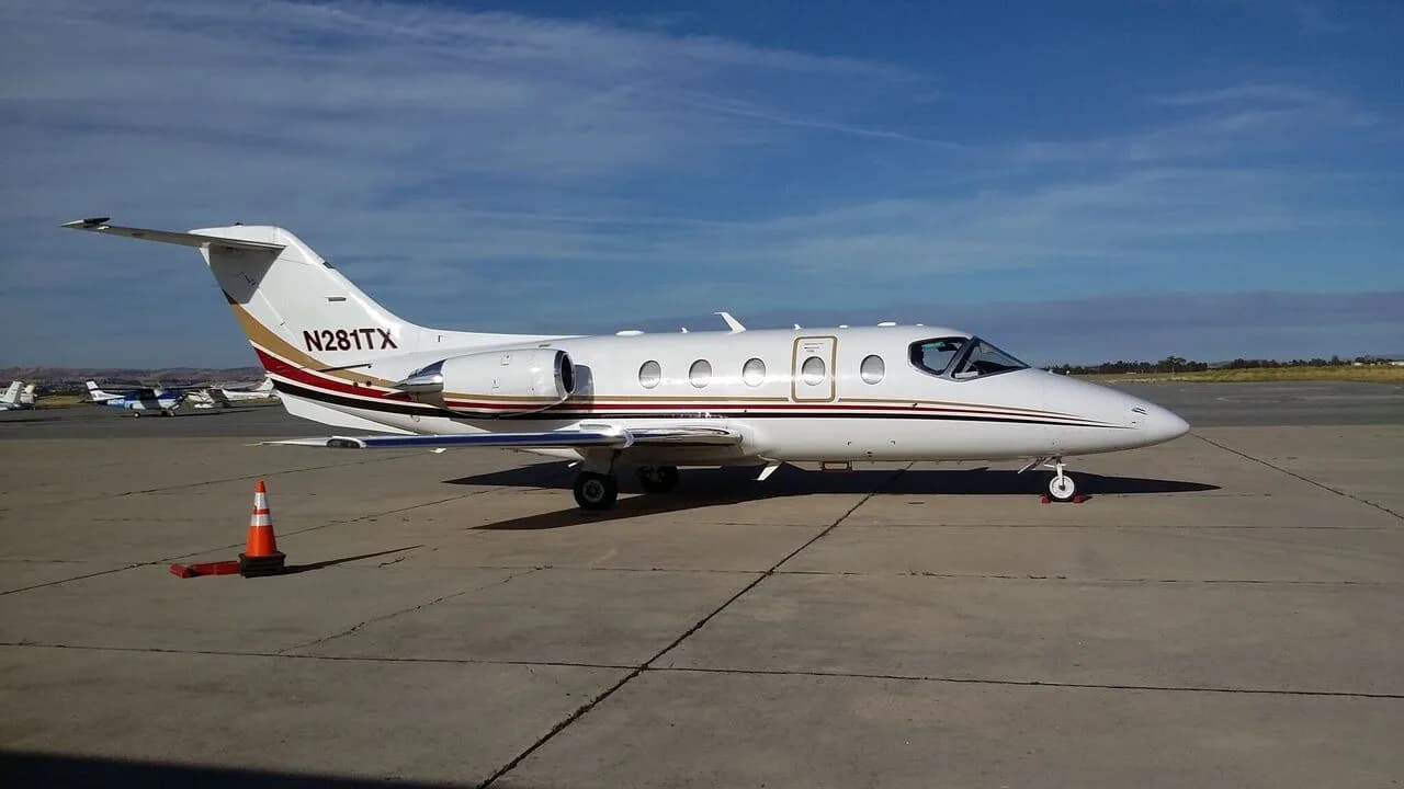 A white Learjet-style private jet with registration N281TX and gold/red stripes parked on an airport tarmac.