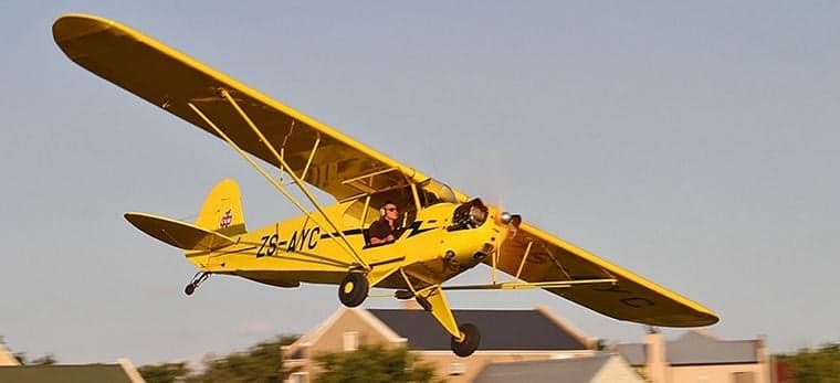 A yellow Piper Cub aircraft is flying over a residential area.
