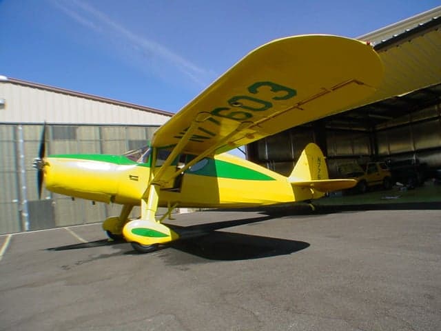 A yellow and green Fairchild 24 aircraft with registration N77603 parked outside a hangar.