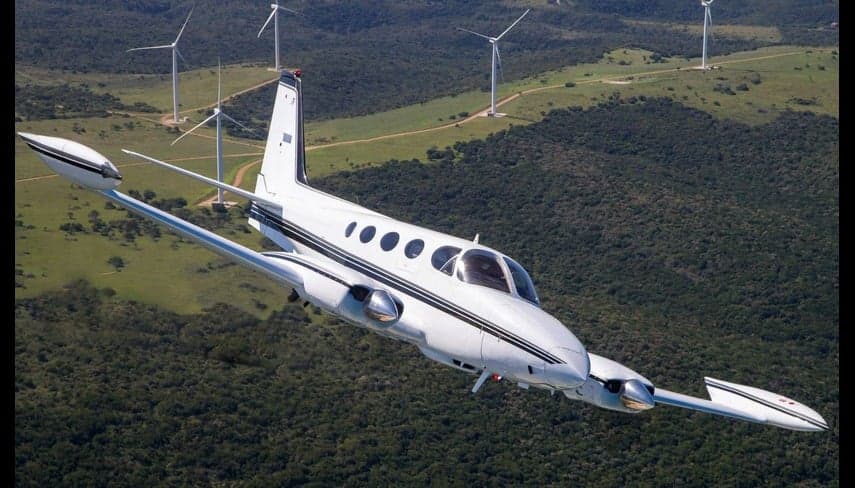A white and silver Beechcraft King Air flying over a green landscape with wind turbines in the background.