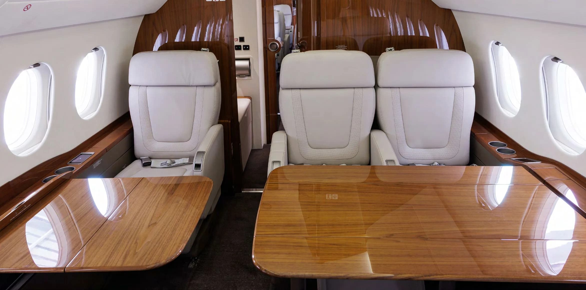 A view inside the business jet cabin showing two sets of light grey leather seats facing each other with polished wooden tables.