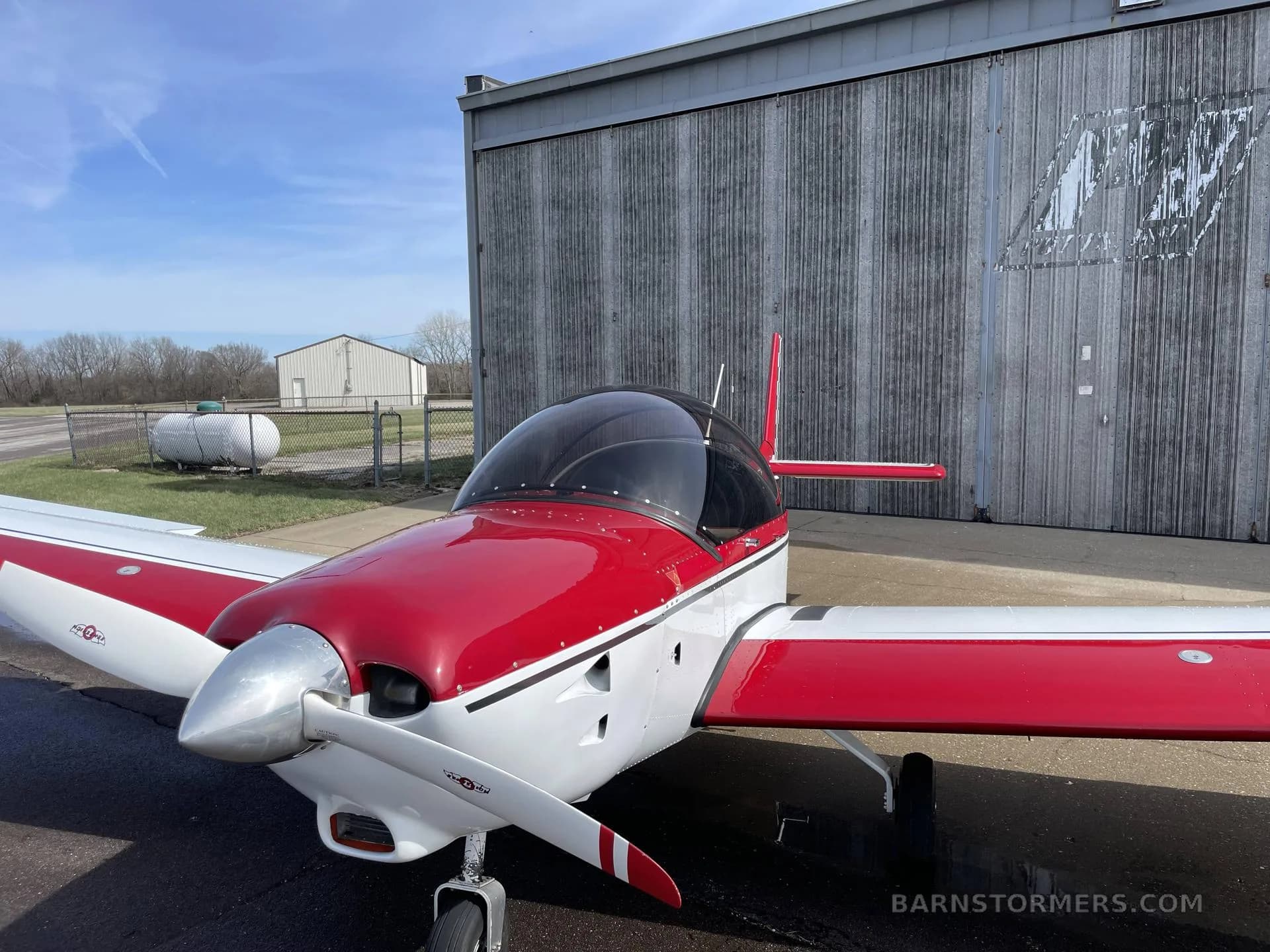 A close-up front-right view of a red and white single-engine aircraft with a silver propeller, parked in front of a hangar.