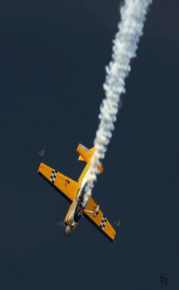 A yellow aerobatic aircraft with checkerboard wings flies inverted, leaving a smoke trail.