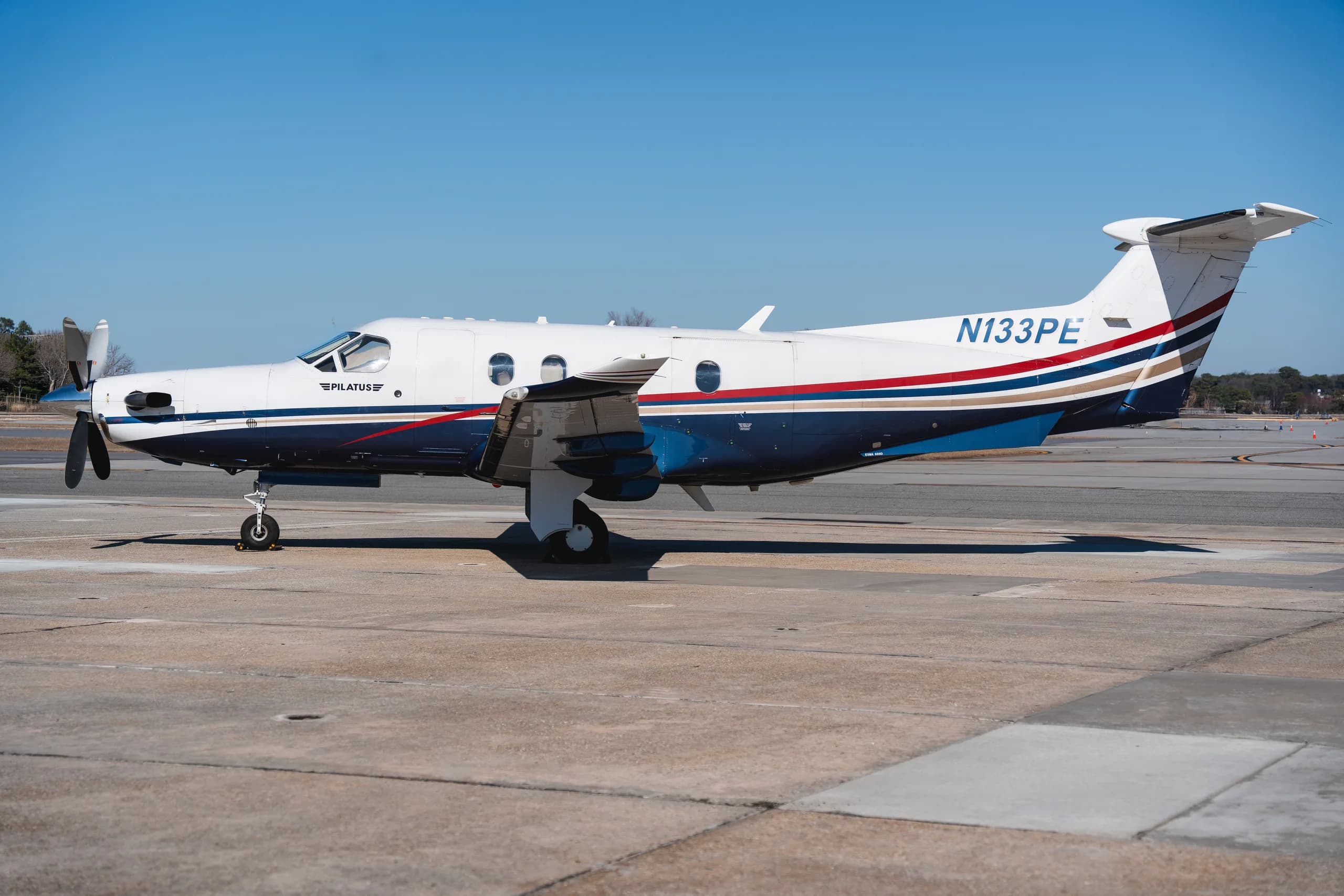A side view of the Pilatus PC-12 turboprop aircraft, registration N133PE, with a white, blue, red, and gold stripe paint scheme, parked on an airfield.