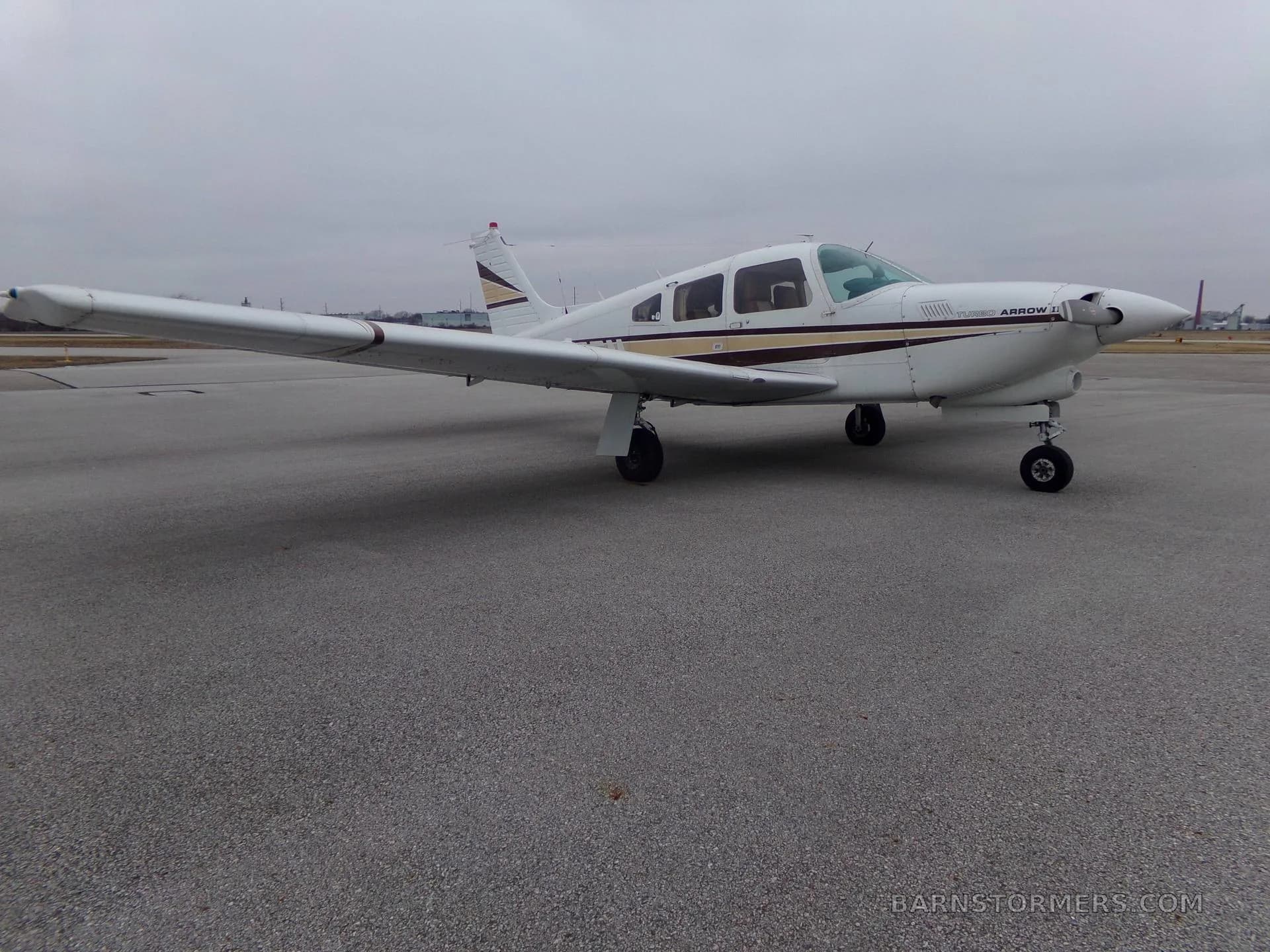 A white Piper Turbo Arrow III aircraft with brown and gold stripes is parked on an asphalt tarmac under an overcast sky, viewed from the front-right.