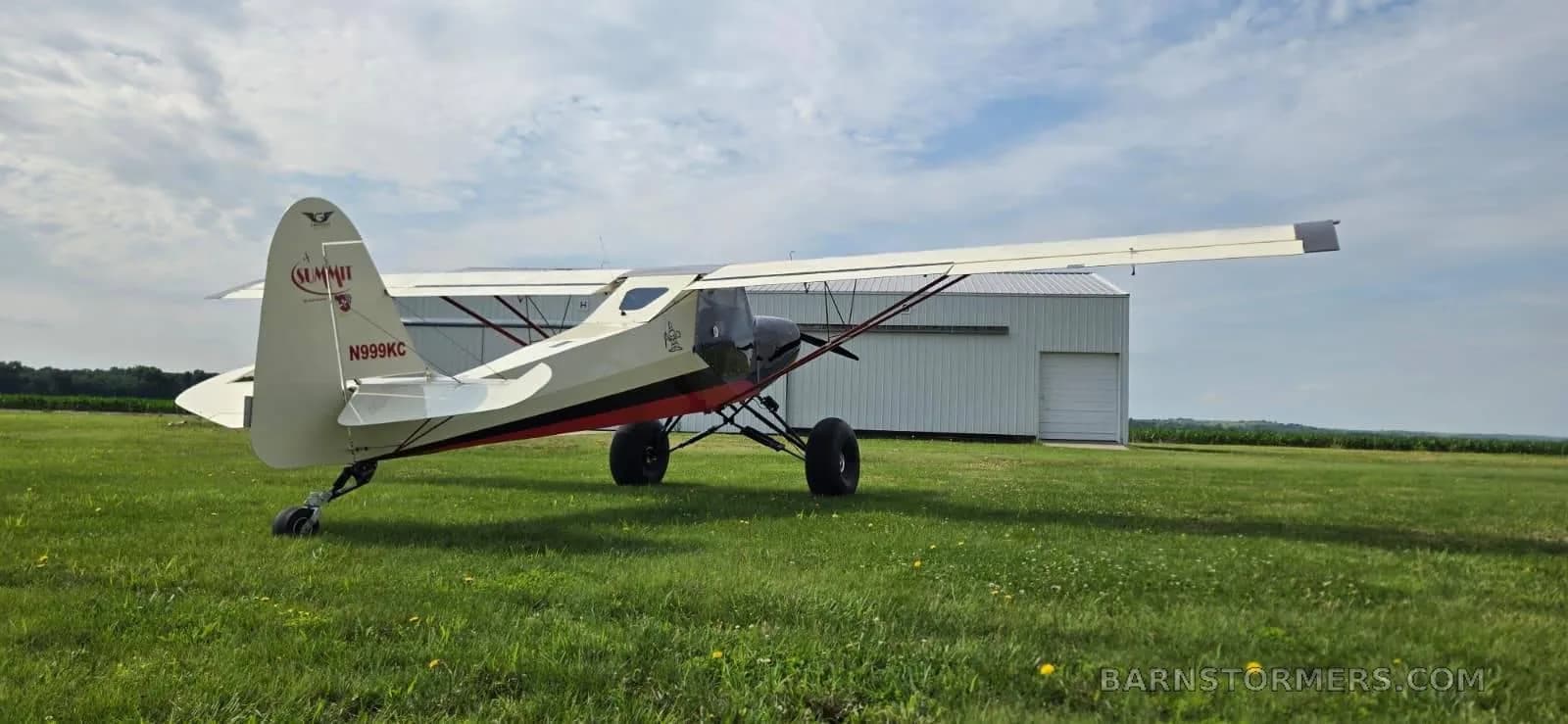 The tail and right side of a cream, red, and black bush plane with large tires are visible, parked on a grassy field near a hangar.
