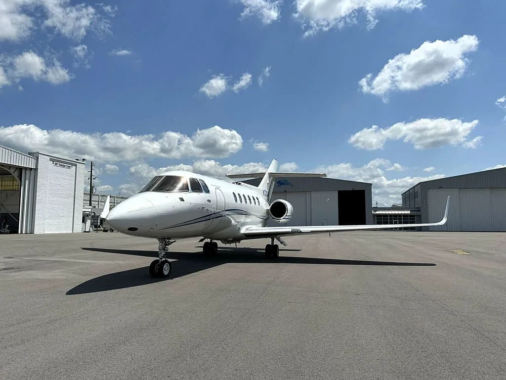 A front-quarter view of a white business jet, N732AA, with blue stripes parked on an tarmac under a blue sky with clouds.