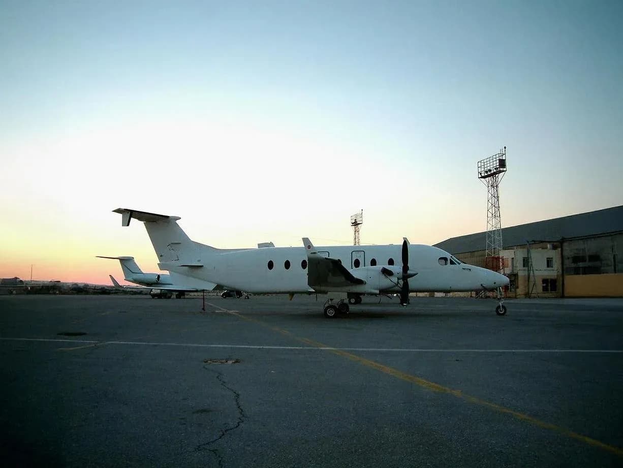 A white twin-engine turboprop aircraft is parked on an airport apron at dawn or dusk, with another aircraft and buildings in the background.