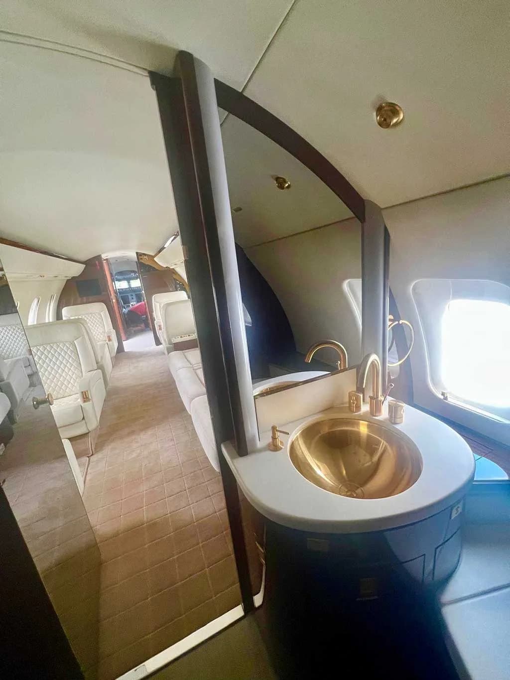 A private jet lavatory with a gold-colored sink, a large mirror, and dark wood cabinetry.