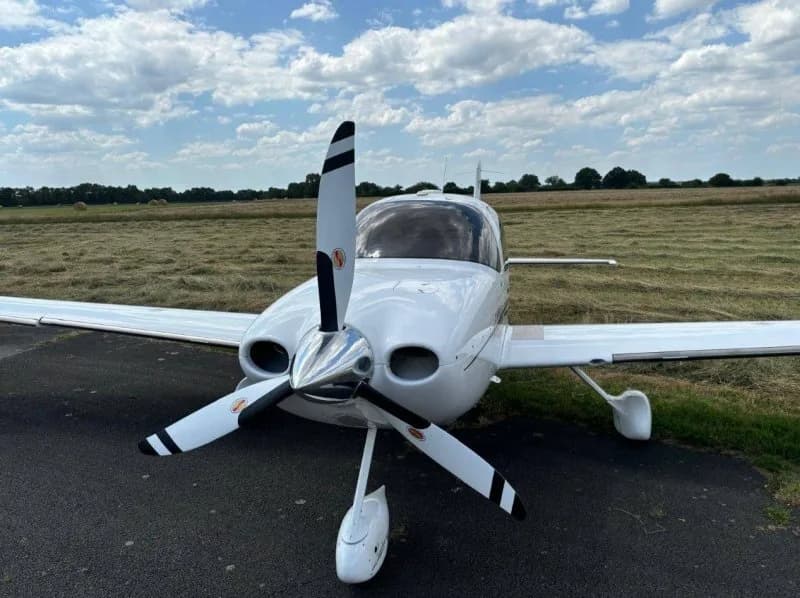 Front view of a white Cirrus SR22 aircraft with a three-bladed propeller on a grassy airfield.