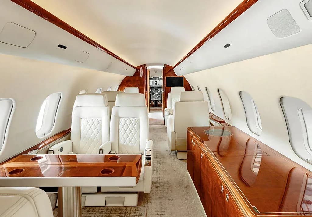 The luxurious interior of a private jet cabin features white quilted leather seats and polished wood tables.