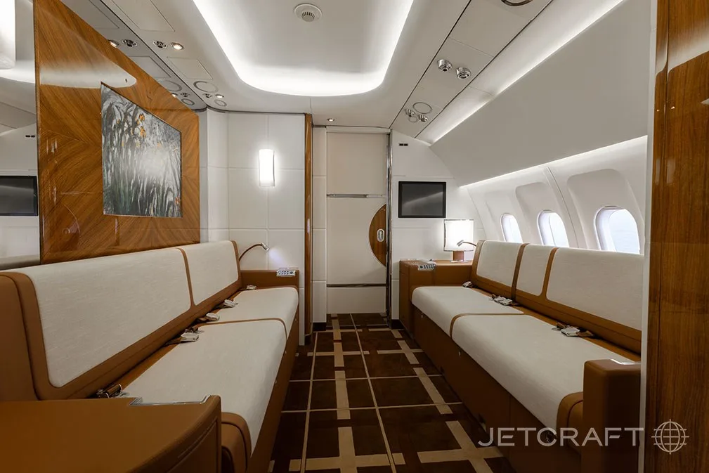 A private aircraft cabin featuring two long cream and brown couches facing each other, with wood paneling and a large screen.