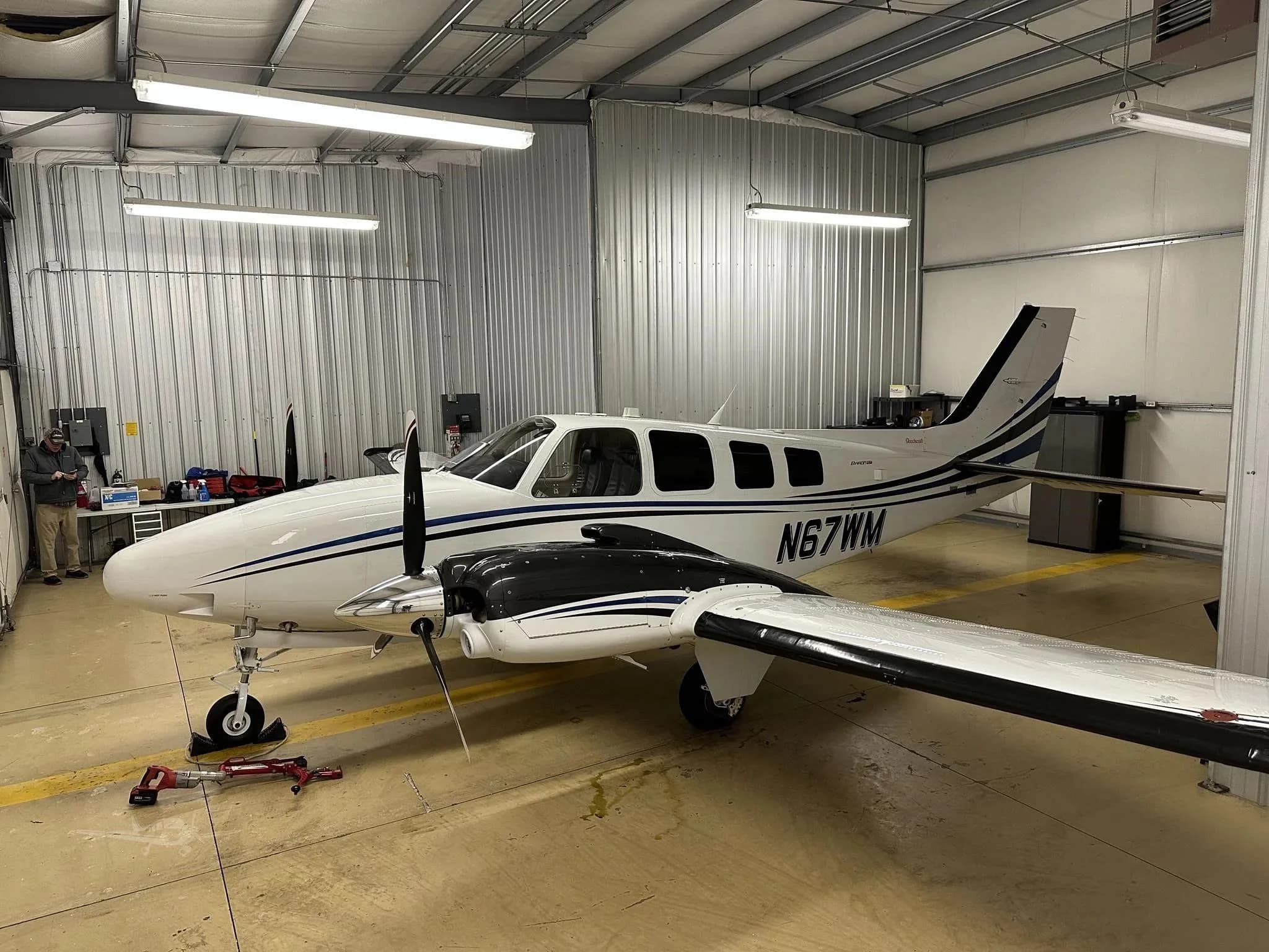 A white and blue Beechcraft Baron N67WM parked inside a hangar, with a person visible on the left.