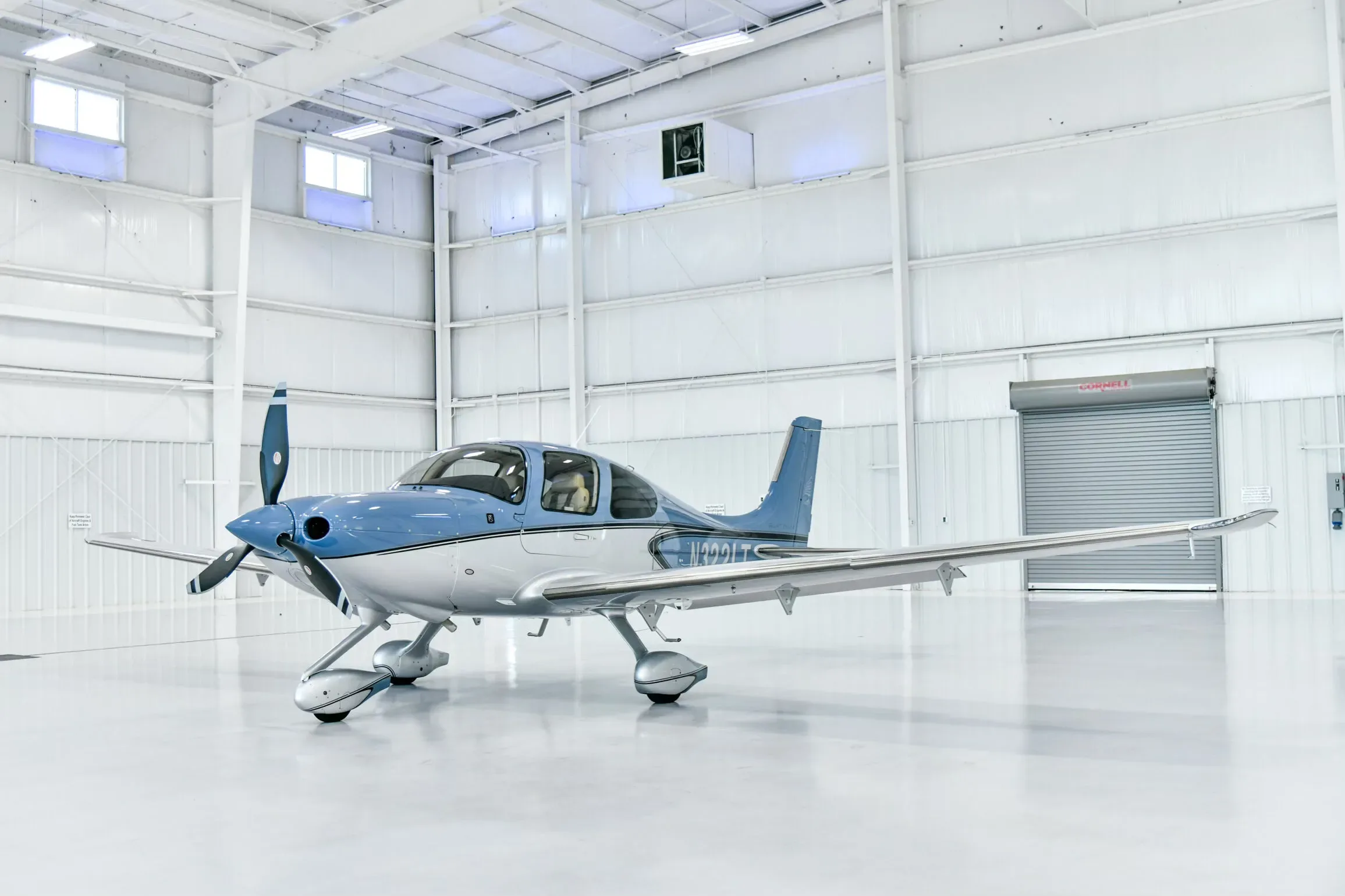 A blue and white Cirrus SR22 aircraft with registration N322LT parked in a brightly lit hangar, viewed from the front quarter.