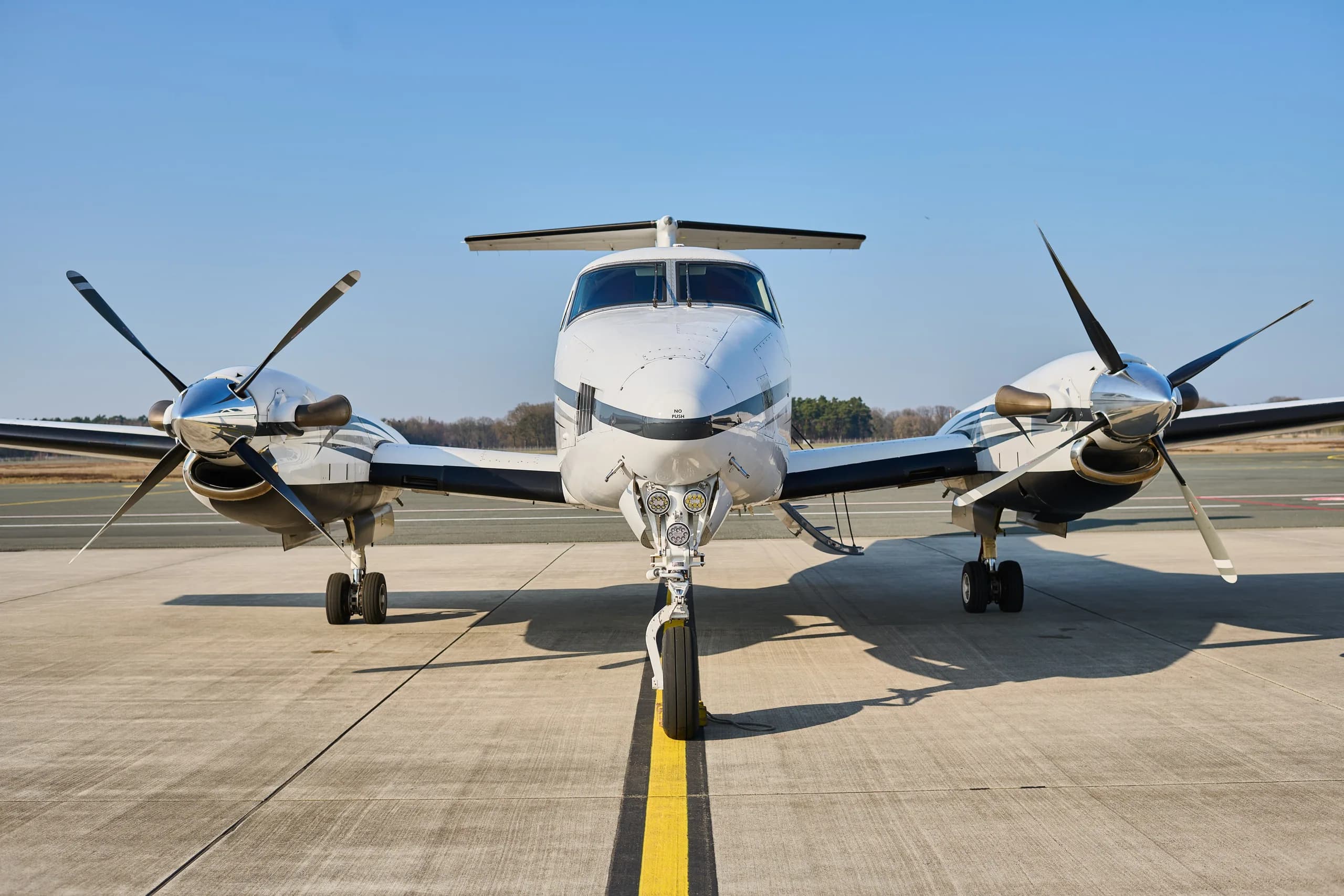 A head-on view of a white and black twin-turboprop aircraft, likely a King Air, parked on an airfield.